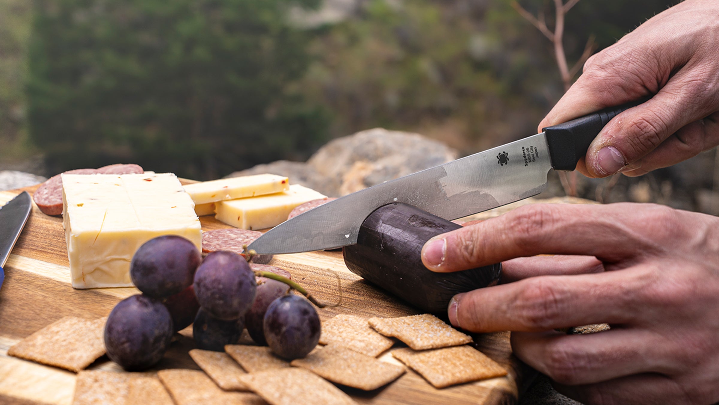 A Spyderco Kitchen knife cutting summer sausage outdoors on a cutting board. 